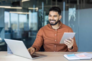 Modern bir ofiste çalışan mutlu profesyonel bir adam laptopu ve tableti arasındaki görevleri dengeliyor, verimliliği, uzaktan çalışmayı ve çağdaş iş iletişimini temsil ediyor.