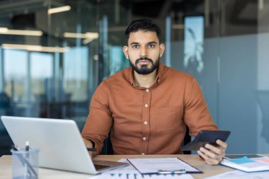 İş adamı, elinde hesap makinesi olan bir dizüstü bilgisayarla çalışırken kameraya bakıyor. Şirket işyerindeki finansal verileri ve belgeleri analiz ediyor.