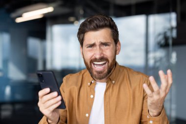 Sinirli ve duygusal bir ifadeyle akıllı telefon tutan genç bir adam, kötü haberlere ya da stresli bir telefon mesajına tepki verirken eliyle işaret ediyor.