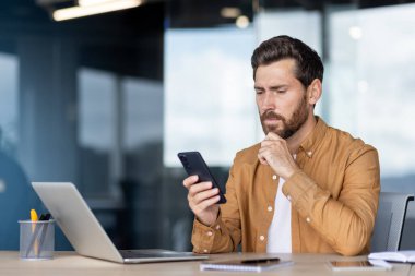 Akıllı giyimli bir iş adamı masasında oturur, akıllı telefonunu incelerken dizüstü bilgisayar ve defter yakınlarda oturur, verileri işler ve iş günü çözümleri planlar.