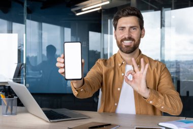 Uygulama ya da web sitesi modeli için beyaz ekranlı bir cep telefonu sunan gülümseyen profesyonel bir adam, ofis masasında dizüstü bilgisayarla otururken iyi bir hareket yapıyor