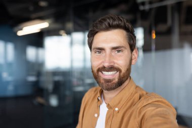 Kameraya gülümseyen sakallı, özgüvenli bir adam, modern bir şirket ofisinde kendi portresini çekiyor, başarıyı, profesyonelliği ve pozitif liderliği ifade ediyor.