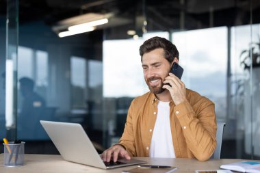 Parlak, modern bir ofis masasında dizüstü bilgisayarda yazarken genç bir profesyonelin telefonunda gülümsemesi, uzaktan çalışma sırasında ya da sanal toplantı sırasında birden fazla görevi güvenle yerine getirmesi, üretken bir çalışma.