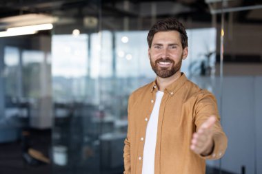 Genç işadamı modern bir ofiste duruyor, kendine güvenerek gülümsüyor ve yeni fırsatları, güveni ve başarılı bir ortaklık kurmayı temsil eden hoş bir tokalaşma teklif ediyor.