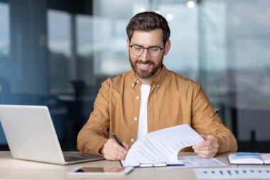 Sakallı ve gözlüklü gülümseyen bir adam ahşap bir masada oturmuş, modern ofis ortamında bir dizüstü bilgisayar üzerinde çalışırken resmi iş belgelerini inceliyor ve imzalıyor.