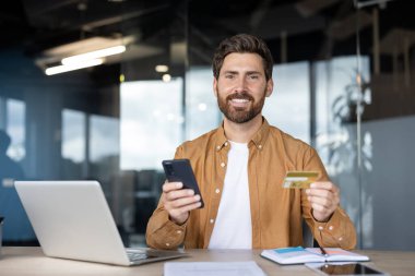 Gülümseyen adam güvenli çevrimiçi alışveriş ve bankacılık işlemleri için cep telefonu ve kredi kartı kullanarak modern ofis ortamından finansmanı dijital olarak yönetiyor.