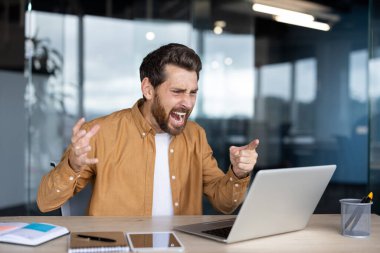 Stresli bir adam dizüstü bilgisayarla çalışırken aşırı hayal kırıklığı ve öfke yaşıyor, ofis ortamında bağırıyor ve el kol hareketi yapıyor, stres, patinaj ve teknik problemleri temsil ediyor.