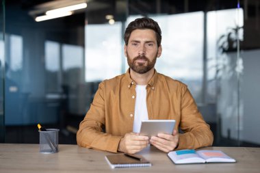 Dijital tableti aktif olarak tutan ve kameraya bakan adam, ofis ortamında modern çalışmayı, teknoloji entegrasyonunu ve profesyonel iletişimi temsil ediyor.