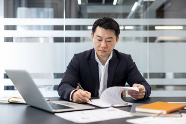 Modern bir ofiste Asyalı işadamı mali raporları inceliyor, sözleşmeleri imzalıyor ve yönetim ve muhasebe görevlerine odaklanmış bir dizüstü bilgisayar üzerinde çalışıyor.