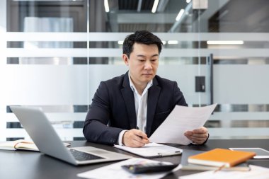 Modern bir masada çalışan Asyalı işadamı, önemli kağıtlara ve finansal verilere odaklanıyor, raporları analiz ediyor ve profesyonel bir ortamda iş görevlerini yönetmek için notlar alıyor.