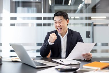 Asyalı işadamı başarısını kutluyor, elinde laptop ve belgelerle masasında oturuyor, ofis ortamında iyi haberler aldıktan sonra sevinç ve zafer bildiriyor.