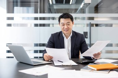 Asyalı işadamı, gülümsüyor ve odaklanıyor, modern ofis masasında önemli mali belgeleri ve raporları inceliyor, başarılı iş operasyonları için veri ve analizleri yönetiyor