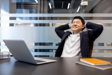 Mutlu Asyalı işadamı, elleri başının arkasında arkasındaydı. Tatmin olmuş gözlerle dizüstü bilgisayarındaki görevlerini tamamladıktan sonra bir anlık rahatlama ve başarının tadını çıkarıyordu.