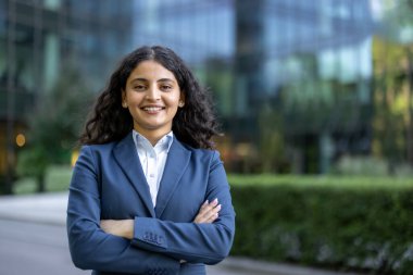 Kendine güvenen genç iş kadını kollarını kavuşturmuş modern bir şirket binasının önünde kameraya gülümsüyor başarılı bir liderlik ve profesyonel yetkiyi temsil ediyor.