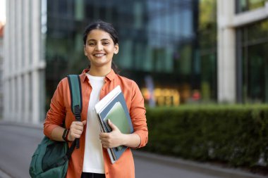 Sırt çantası takan ve kitap tutan Hintli genç bir kız öğrenci, modern bir üniversite kampüsünde eğitim, öğrenme ve öğrenci hayatını somutlaştırırken kendine güvenerek gülümsüyor.