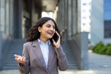 Akıllı telefonuyla neşeli bir konuşma yaparken gülümseyen ve el kol hareketi yapan profesyonel genç bir kadın, modern ofis arkaplanına karşı güvenli bir şekilde dışarıda yürüyor.