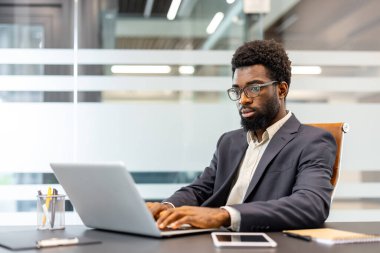 Profesyonel bir adam gözlük ve takım elbise takıyor, bir masada oturuyor ve bir dizüstü bilgisayarda daktilo yazıyor, çağdaş bir çalışma alanında işine odaklandığını ve kendini adadığını gösteriyor.