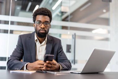 Masa başında oturan takım elbiseli ve gözlüklü profesyonel bir adam elinde akıllı bir telefon, yanında dizüstü bilgisayar ve defterle parlak bir ofis ortamında ileriye bakıyor.