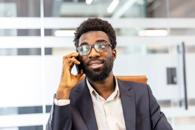 Modern bir ofiste genç bir işadamı cep telefonuyla konuşuyor, akıllı günlük kıyafetlerini giyer, planlama stratejisi ve ağ kurma konusunda kendinden emin bir şekilde kafasını çeviriyor.