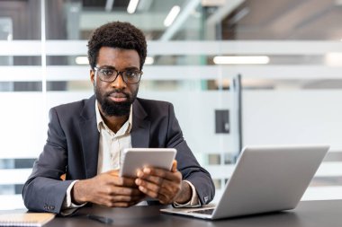 Takım elbiseli ve gözlüklü kendine güvenen profesyonel bir adam bir ofis ortamında tablet tutarken ileriye bakıyor ve modern iş iletişimini gösteriyor.