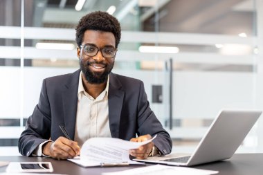 Ofis masasında oturan genç işadamı, kameraya gülümserken bir belgeye, yakınlarda dizüstü bilgisayarı ve akıllı telefonuyla, başarı ve anlaşmayı temsil ediyor.