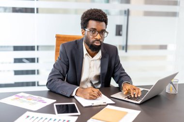 Profesyonel işadamı, ofis masasında özenle çalışırken, aynı anda bir dizüstü bilgisayarda notlara odaklanarak, kurumsal bir ortamda üretkenlik ve çoklu görev göstererek,