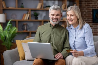Üst düzey bir çift evde bir kanepede dinleniyor, elinde kredi kartı tutarken gülümsüyor ve dizüstü bilgisayara bakıyor, güvenli bir şekilde online alımlar yapıyor ve internet üzerinden finansman yönetiyor.