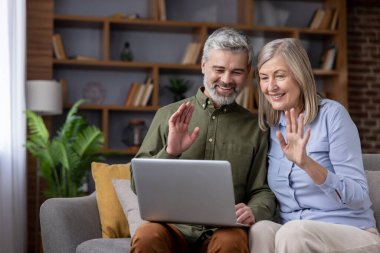 Olgun bir çift evdeki koltukta dinleniyor, video görüşmesi sırasında gülümsüyor ve dizüstü bilgisayara el sallıyor, modern teknoloji sayesinde uzak aile veya arkadaşlarıyla aktif olarak iletişim kuruyor.