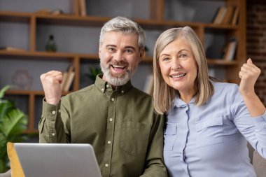 Mutlu, olgun bir çift kanepeye oturup kameraya bakıyor ve yumruklarını başarıyla kaldırıyor. Ev ortamında dizüstü bilgisayar kullanırken heyecan ve başarı gösteriyorlar.