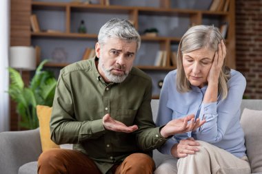 Olgun yaşlı çift, evde bir kanepede oturup duygusal stres ve zorluk yaşarken, erkek hayal kırıklığını ifade ederken kadın üzgün ve üzgün görünüyor.