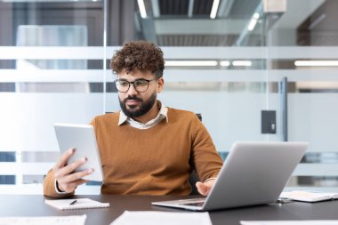 Genç işadamı, üretken bir ortamda veriyi ve dijital bağlantıyı gösteren modern bir şirket masasında çalışırken tablet üzerindeki verileri analiz ediyor.