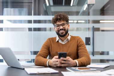 Kıvırcık saçlı ve gözlüklü gülümseyen genç adam izleyiciye bakıyor. Sırasında dizüstü bilgisayar ve modern ofis ortamında belgelerle otururken akıllı telefon kullanıyor.