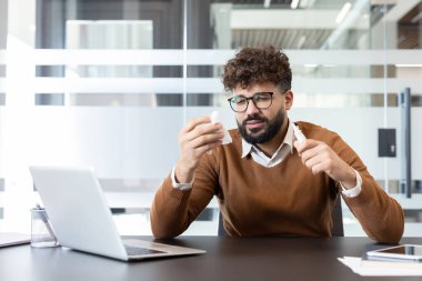 Masadaki genç erkek ofis çalışanı, elinde burun ve doku akıntısı, elinde burun spreyi tutarken tıkanmış ve dikkati dağılmış bir şekilde dizüstü bilgisayar üzerinde çalışırken kendini kötü hissediyor.