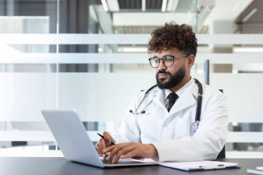 Genç erkek doktor bir dizüstü bilgisayar üzerinde çalışıyor, çevrimiçi istişareler sunuyor ve çağdaş bir sağlık ofisinde hasta kayıtlarını yönetiyor, dijital sağlık ve profesyonel yardımı sembolize ediyor.