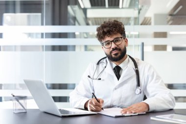 Beyaz önlüklü ve steteskoplu genç bir erkek doktor, modern bir klinikte dizüstü bilgisayarla bir masaya notlar yazarken güvenle gülümsüyor.