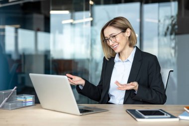 Gözlüklü genç iş kadını aktif olarak online bir video görüşmesine katılıyor ya da bilgisayarını kullanıyor, bir şirket ofisinden sunuyor ve gülümsüyor.