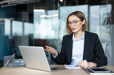 Gözlüklü genç bir iş kadını, modern bir şirket ofisinden bilgisayarına uzaktan kumandalı bir toplantı düzenliyor, online iletişim kurarken aktif olarak tartışıyor ve not alıyor.