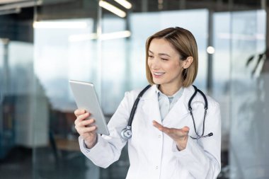 Beyaz önlük ve steteskop giyen genç bir kadın doktor, dijital tabletle yapılan teletıp görüntülerinde gülümseyip el kol hareketi yapıyor ve uzaktan sağlık desteği teklif ediyor.