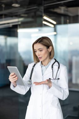 Sağlık verilerini sunmak ve tartışmak için dijital tablet kullanan beyaz önlüklü kadın doktor, bir klinik ortamında modern tıp ve teknolojiyi gösteriyor