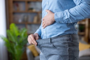 Mavi gömlek giyen kişi gururla kilo kaybettiğini gösteren bol kot pantolon giyer, sağlık gelişimini, diyet bağlılığını ve pozitif yaşam tarzını sembolize eder.