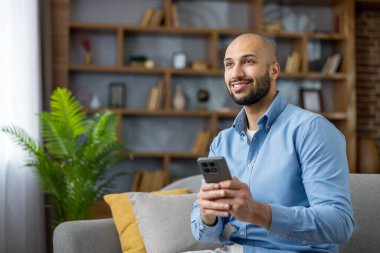 Genç adam gülümsüyor ve rahat bir şekilde kanepede oturuyor, akıllı telefonunu sosyal medyada gezinmek ve rahat oturma odasından internete bağlanmak için kullanıyor.