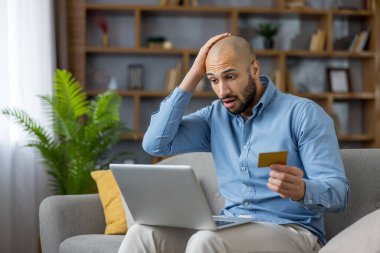 Genç bir adam altın kredi kartını tutuyor ve elini başına koyuyor. Evde dizüstü bilgisayar kullanarak finansal bir sorun ya da online dolandırıcılık yaşarken şok olmuş ve stresli görünüyor.