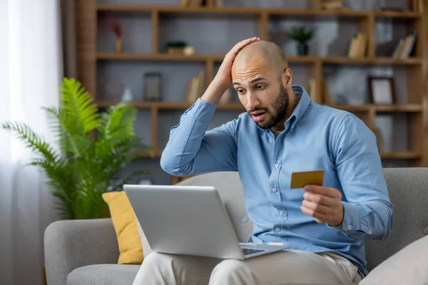 Genç bir adam altın kredi kartını tutuyor ve elini başına koyuyor. Evde dizüstü bilgisayar kullanarak finansal bir sorun ya da online dolandırıcılık yaşarken şok olmuş ve stresli görünüyor.