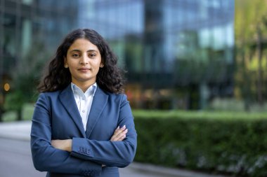 Kendine güvenen, mavi takım elbiseli Hintli bir iş kadını şehrin dışında, kollarını kavuşturmuş, liderlik ve ihtiraslı bir yönetici portresi çiziyor.