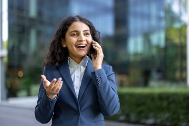 Bulaşıcı bir gülümsemesi olan genç bir iş kadını açık havada canlı bir telefon görüşmesi yapıyor, modern bir şirket geçmişine karşı profesyonel başarının ve bağlantının tadını çıkarıyor.
