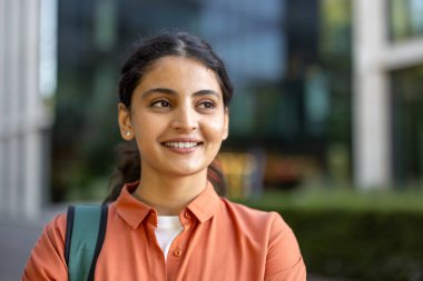 Sırtında sırt çantası ve turuncu bir tişörtle kendinden emin bir şekilde gülümseyen genç bir kız öğrenci üniversite kampüsünün dışında duruyor ve geleceği hakkında iyimser hissediyor.