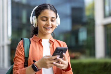 Kampüste yürürken gülümseyen genç kız öğrenci, cep telefonu ve kablosuz kulaklık kullanarak, bir şehir ortamında eğitim, müzik ve dijital bağlantıyı temsil ederek,