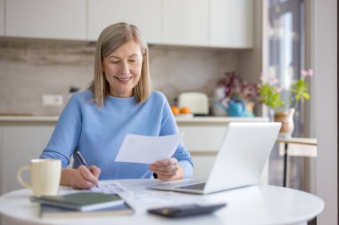 Mutfak masasındaki kıdemli kadın finansal belgeleri analiz ediyor ve dizüstü bilgisayar kullanıyor, ev bütçesini mutlulukla dengeliyor ve gelecekteki harcamalarını kolaylıkla ve güvenle planlıyor.