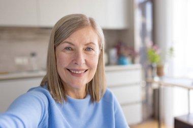 Yüzünde samimi bir gülümsemeyle, mutlu bir emekliliği kucaklayan ve mutlu bir şekilde selfie çeken son sınıf öğrencisi bir kadın. Parlak, modern mutfağında sıcak bir bağ kuruyor.