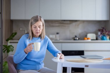Düşünceli son sınıf öğrencisi kadın mutfak masasında oturuyor, elinde bir fincan kahve tutuyor ve evdeki sakin sabah rutini sırasında yaşadığı üzüntü, yalnızlık ve tefekkür hislerini yansıtıyor.